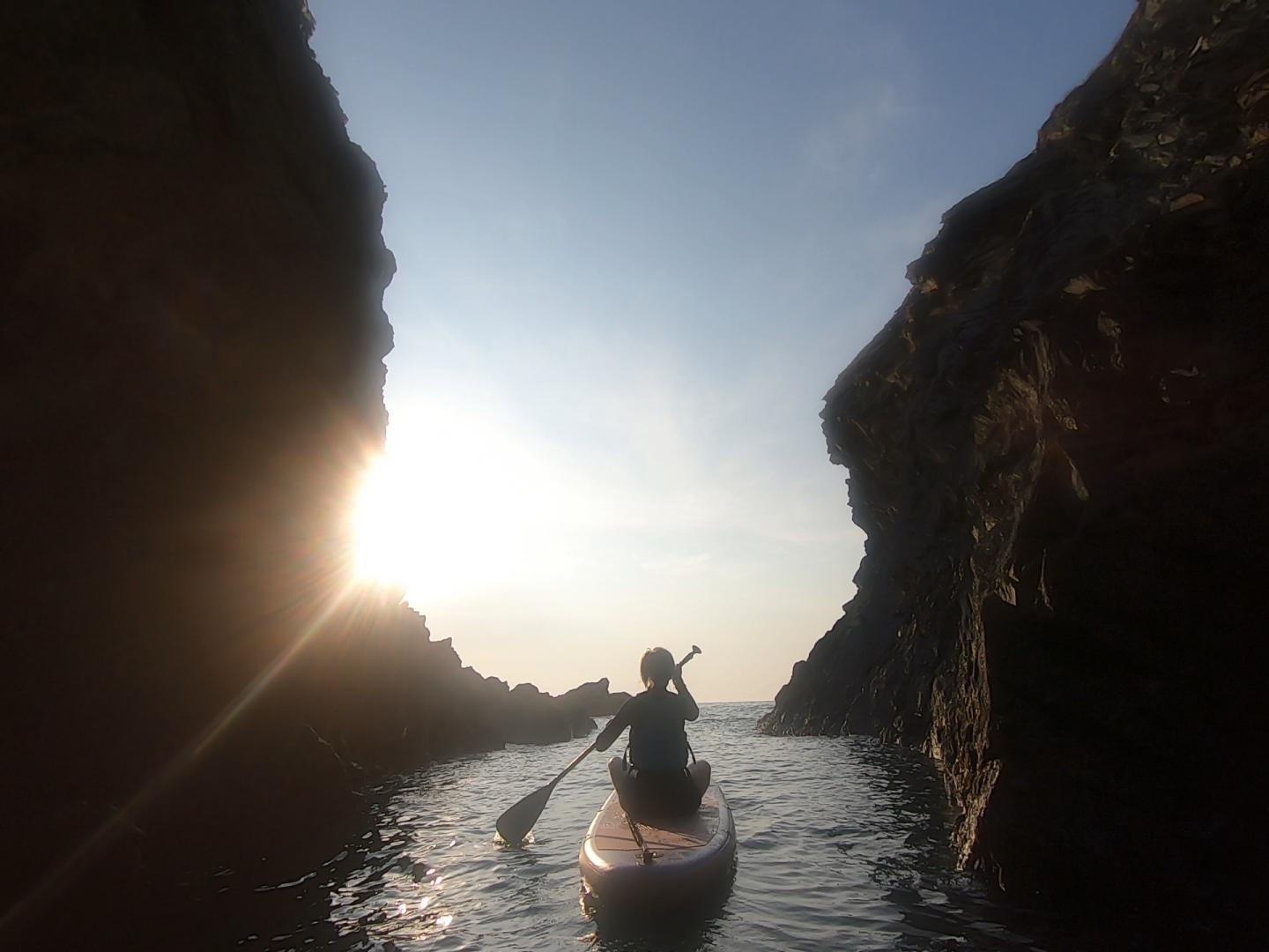 A paddleboarder gliding between towering cliffs toward the low evening sun at the mouth of the Witches Cauldron, Pembrokeshire.